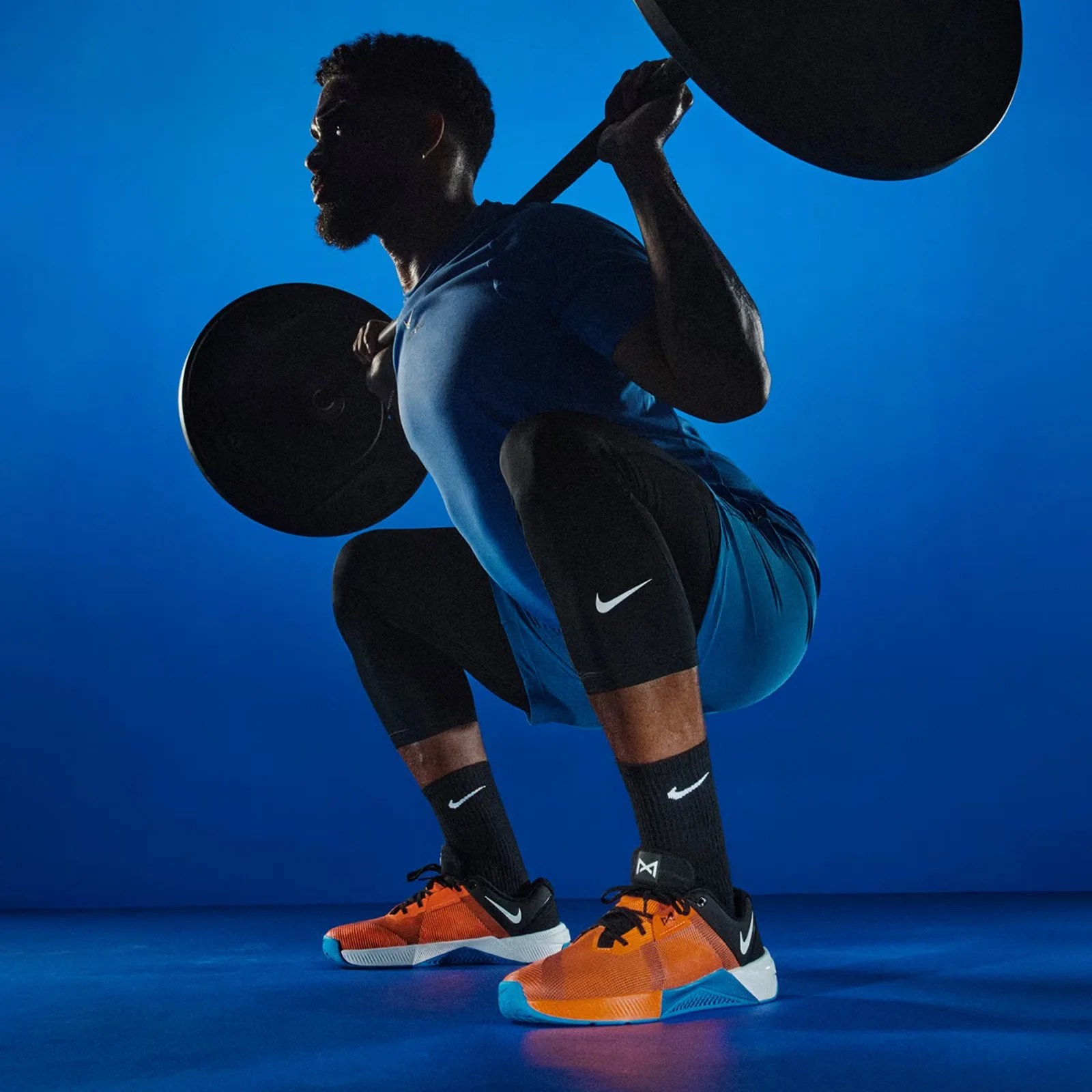A man in Nike athletic apparel and a pair of Nike Metcon 10 gym shoes shown performing a squat with a barbell on his shoulders in a dramatically lit blue room.