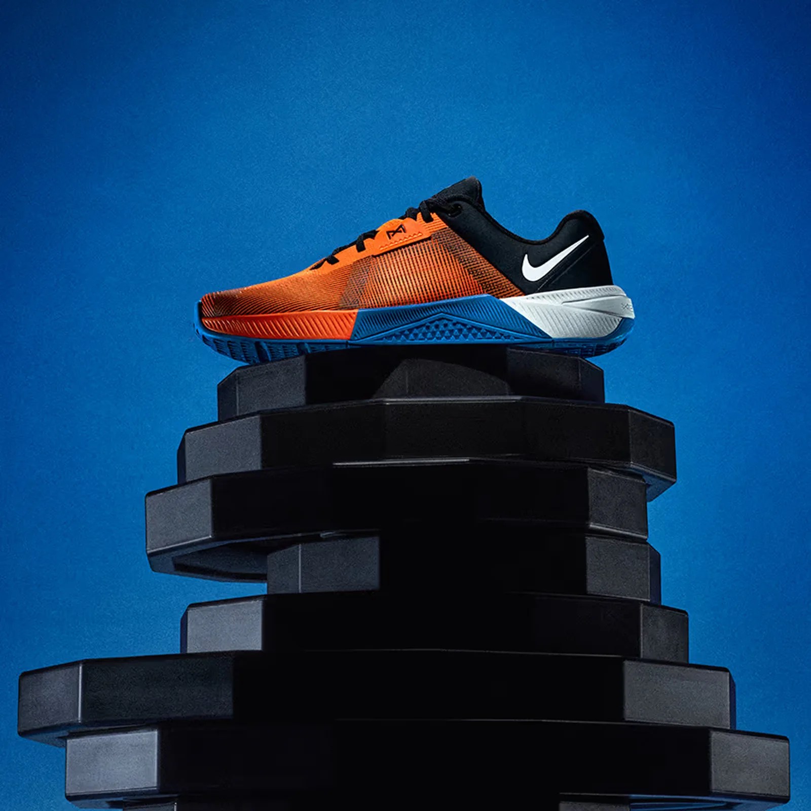 A single Nike Metcon 10 sneaker in blue and orange is shown sitting on top of a pedestal of metal weight plates against a bright blue background. 